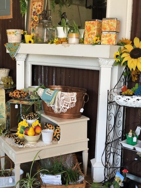 An antique white fireplace mantel and hearth are styled with a rustic copper tub, sunflowers, floral tins, and vintage accents, creating a charming display found in antique stores in Coffeyville, KS.